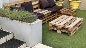 A wooden bench with cushions, a planter with purple flowers, and a yellow pot on a grassy surface. - Coogee Beachside - Hostel (Coogee)