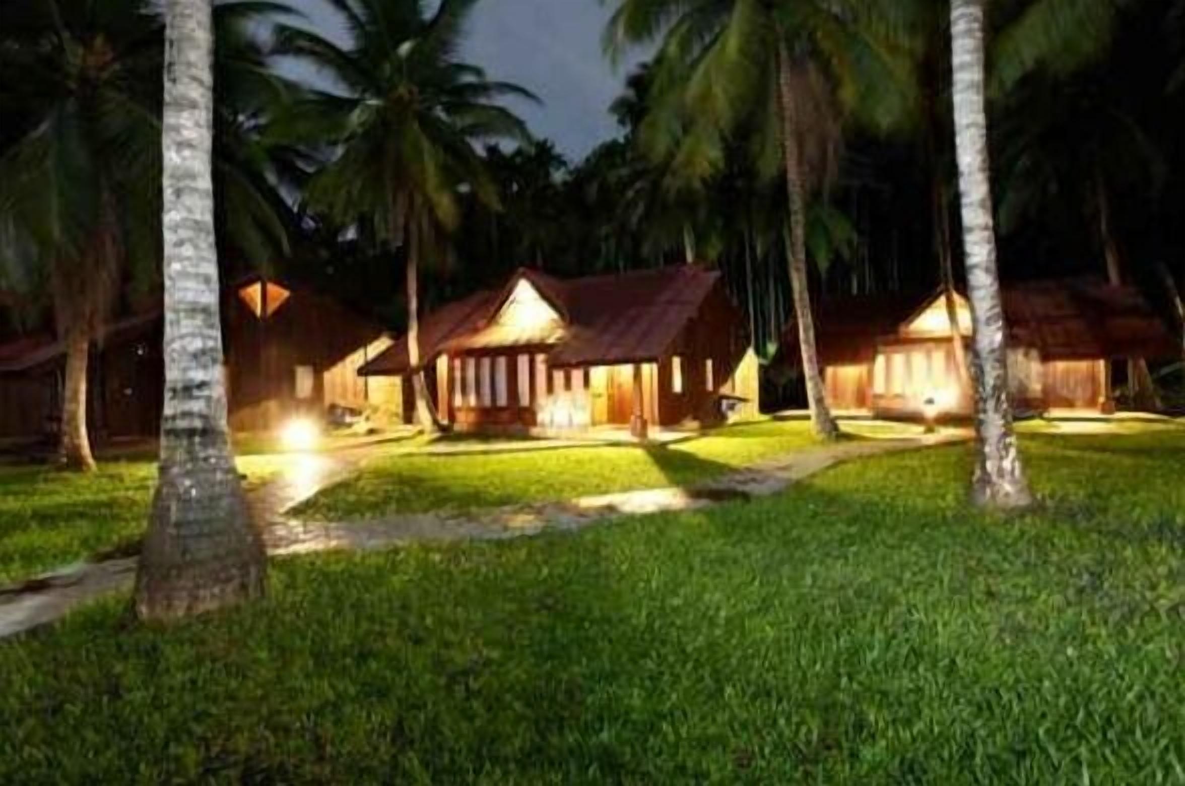 A row of wooden cabins with thatched roofs, surrounded by palm trees and well-maintained grass.