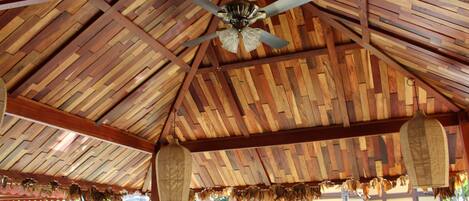 A wooden pavilion with a ceiling fan, thatched roof decorations, and a view of a building in the background.