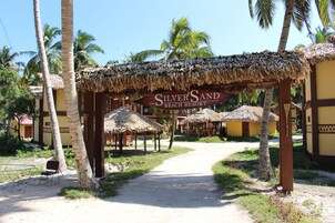 Un sendero que conduce a un resort llamado 'Silver Sand Beach Resort', con edificaciones de techo de paja y palmeras.