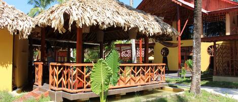A traditional thatched-roof structure with wooden railings, a small water feature, and a palm tree in the foreground.