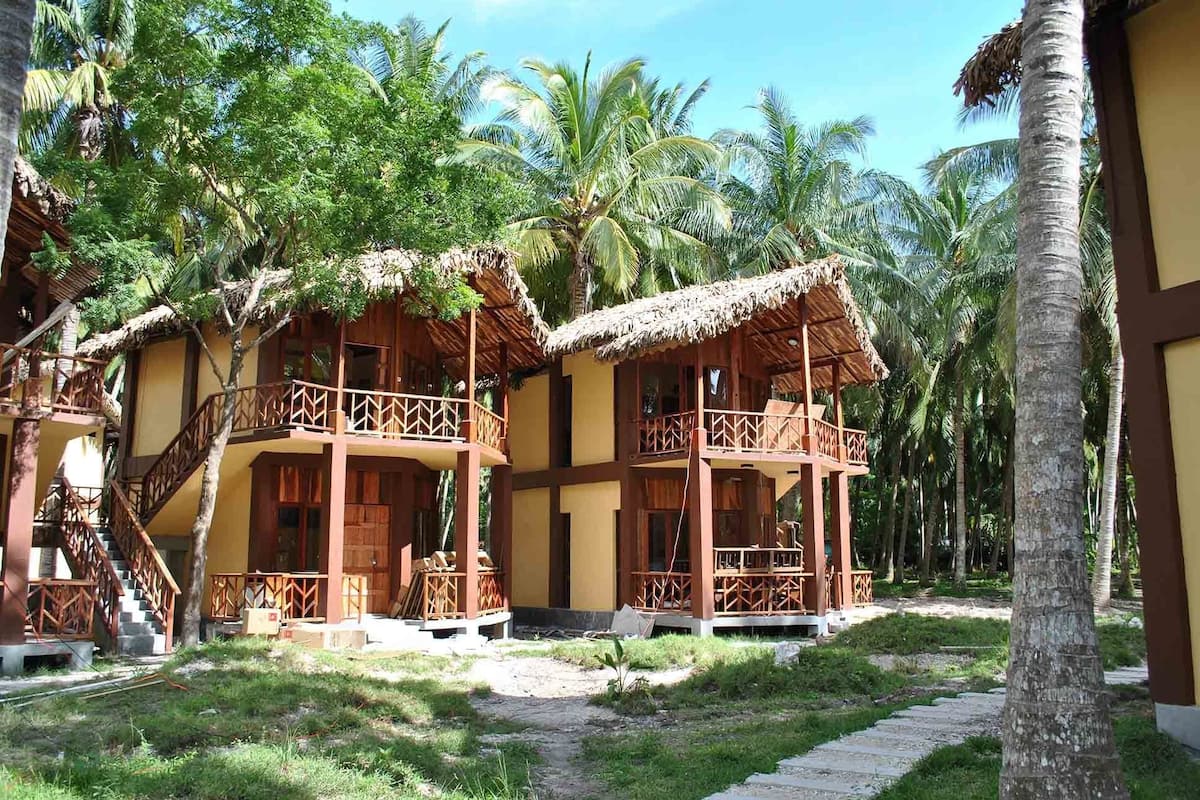 A resort with two wooden bungalows with thatched roofs, surrounded by palm trees.