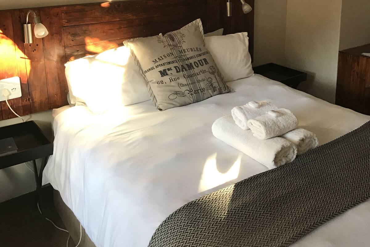 A neatly made bed with white linens, a pillow with text, and a blanket. There are framed pictures on the wall and a lamp on the bedside table.