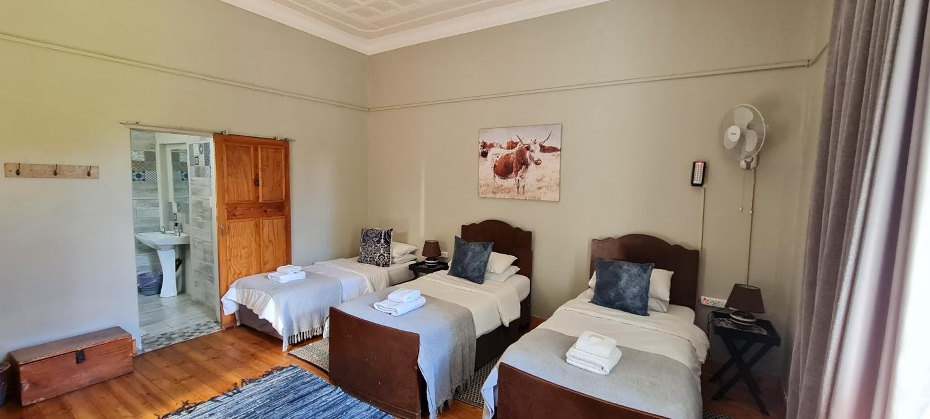 Quarto com duas camas, um guarda-roupa de madeira, um banheiro visível por uma porta de vidro, um ventilador e um quadro de uma vaca.