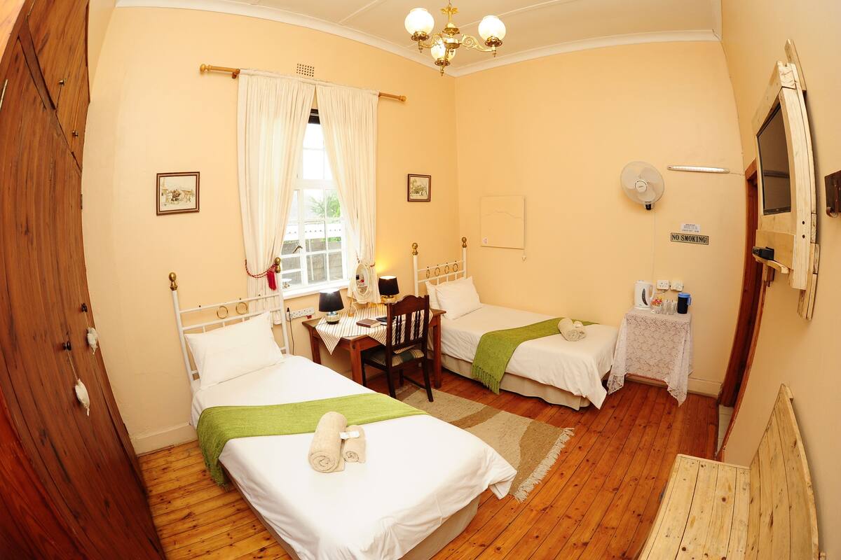 A room with two single beds, a wooden desk with a chair, a small table with a lamp, and a window with curtains.