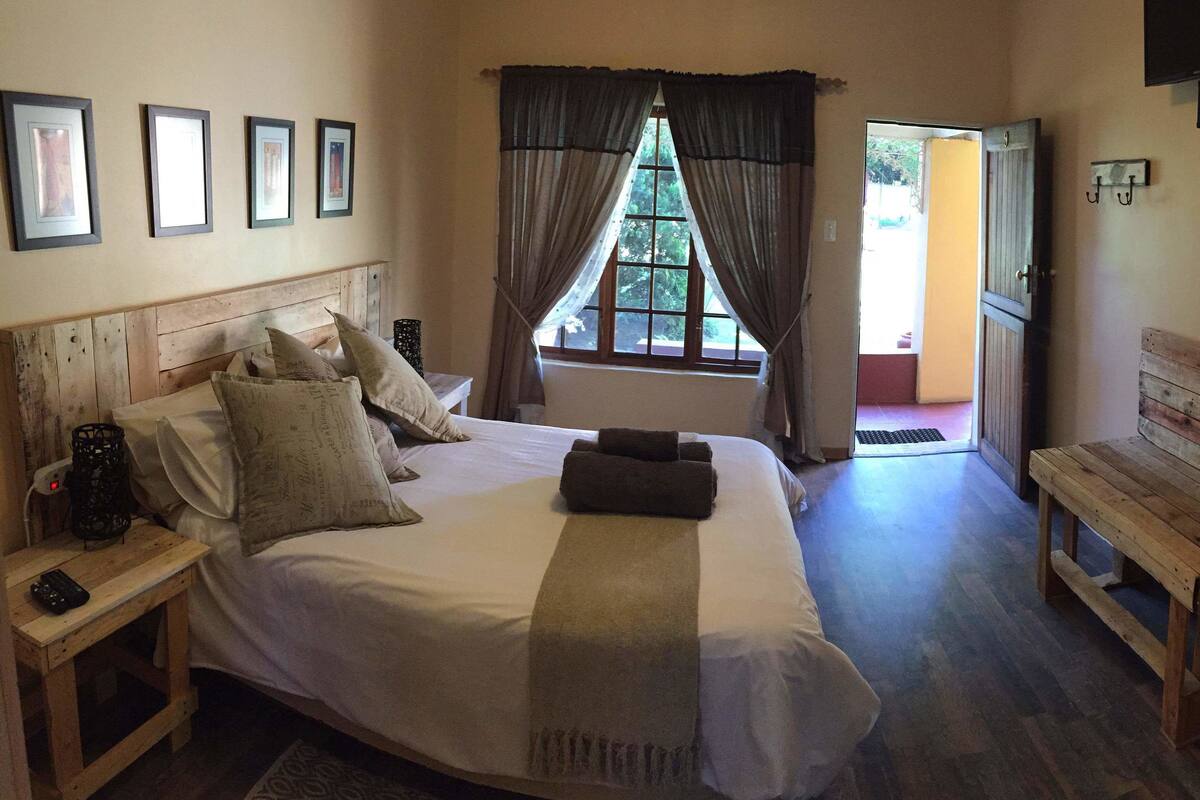 A bedroom with a large bed, a wooden bench, a bedside table, a TV mounted on the wall, and a window with curtains.