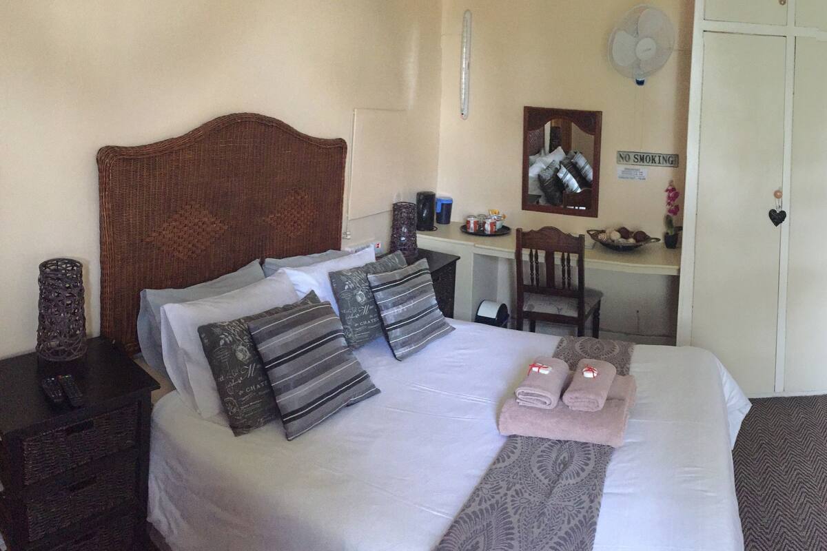 A neatly made bed with pillows, a bedside table with a remote control, a desk with a chair, a mirror, and a wardrobe.