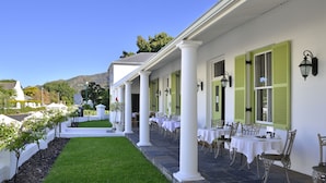 Un área de comedor al aire libre con techo, columnas blancas y contraventanas verdes, mesas dispuestas para comer y un césped cuidado.