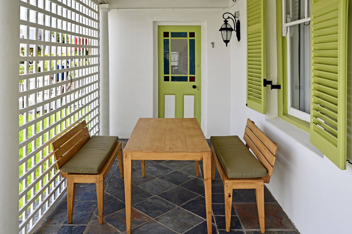 Una mesa de madera con dos bancos, una puerta verde y una pared blanca con una ventana y una lámpara fijada a la pared.
