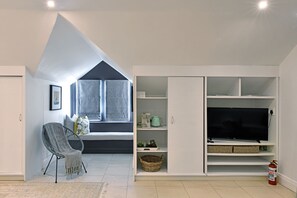 A modern living room with a built-in TV, a grey chair, a window with curtains, and a white shelving unit.