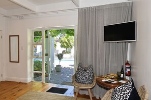 A room with a wooden floor, a chair with leopard print cushions, a small table with a bottle, glasses, and papers, a fire extinguisher, and a TV on the wall.