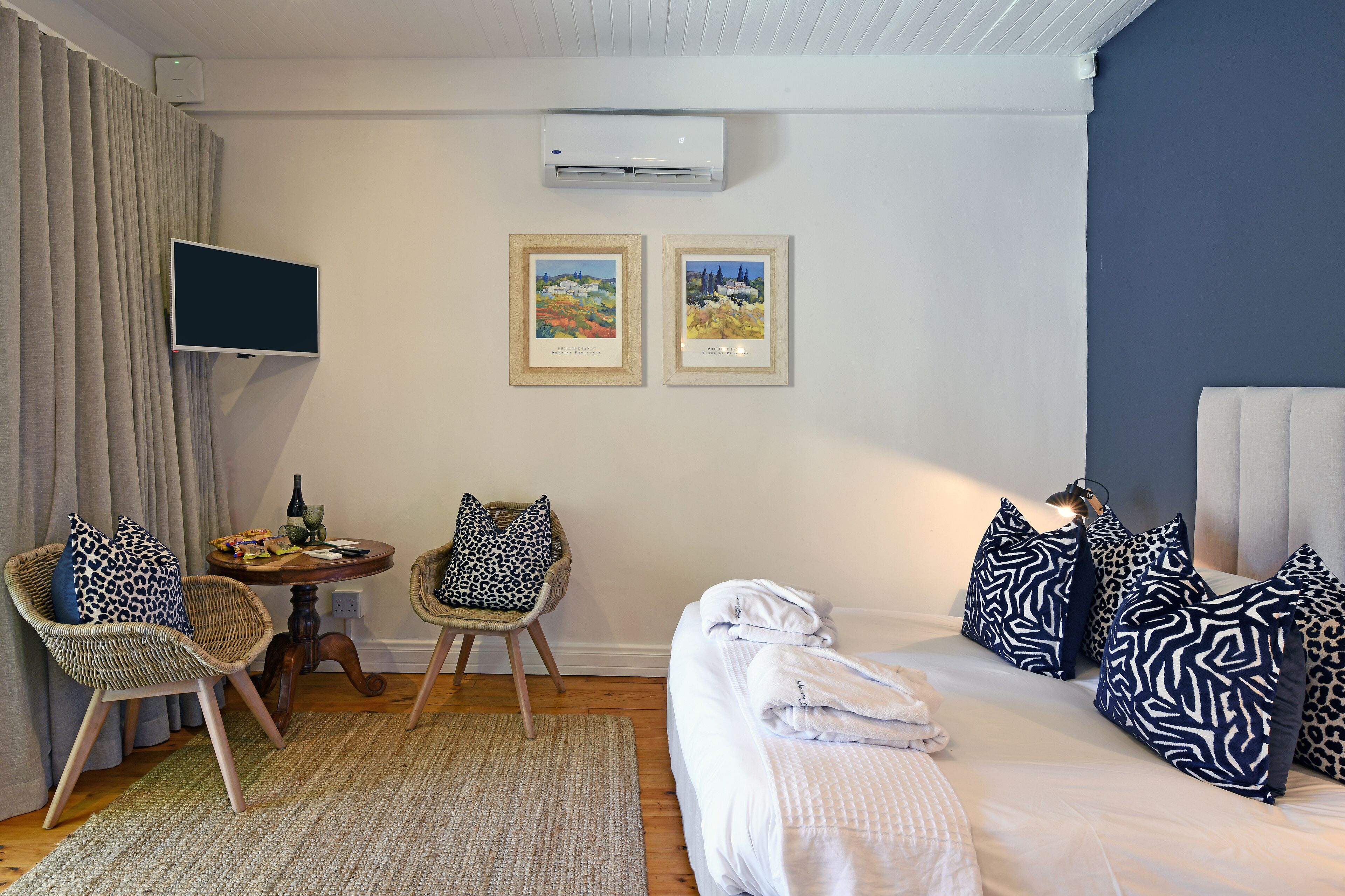 Ett hotellrum med en säng, två stolar, ett litet bord med en flaska och några snacks, två inramade tavlor på väggen och en luftkonditioneringsanläggning.