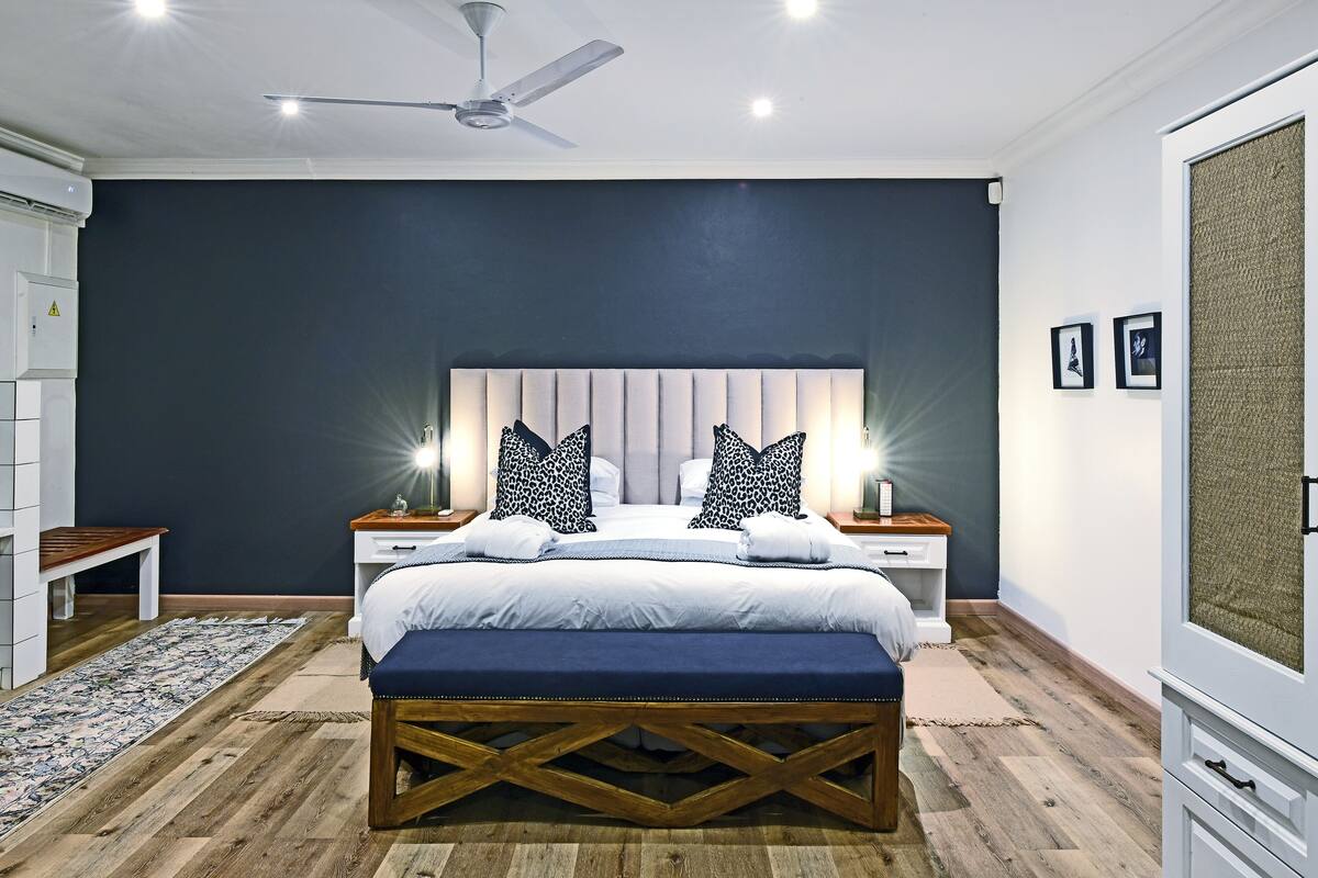 Un dormitorio con cama, mesitas de noche, un banco, ventilador de techo y un ventanal grande.