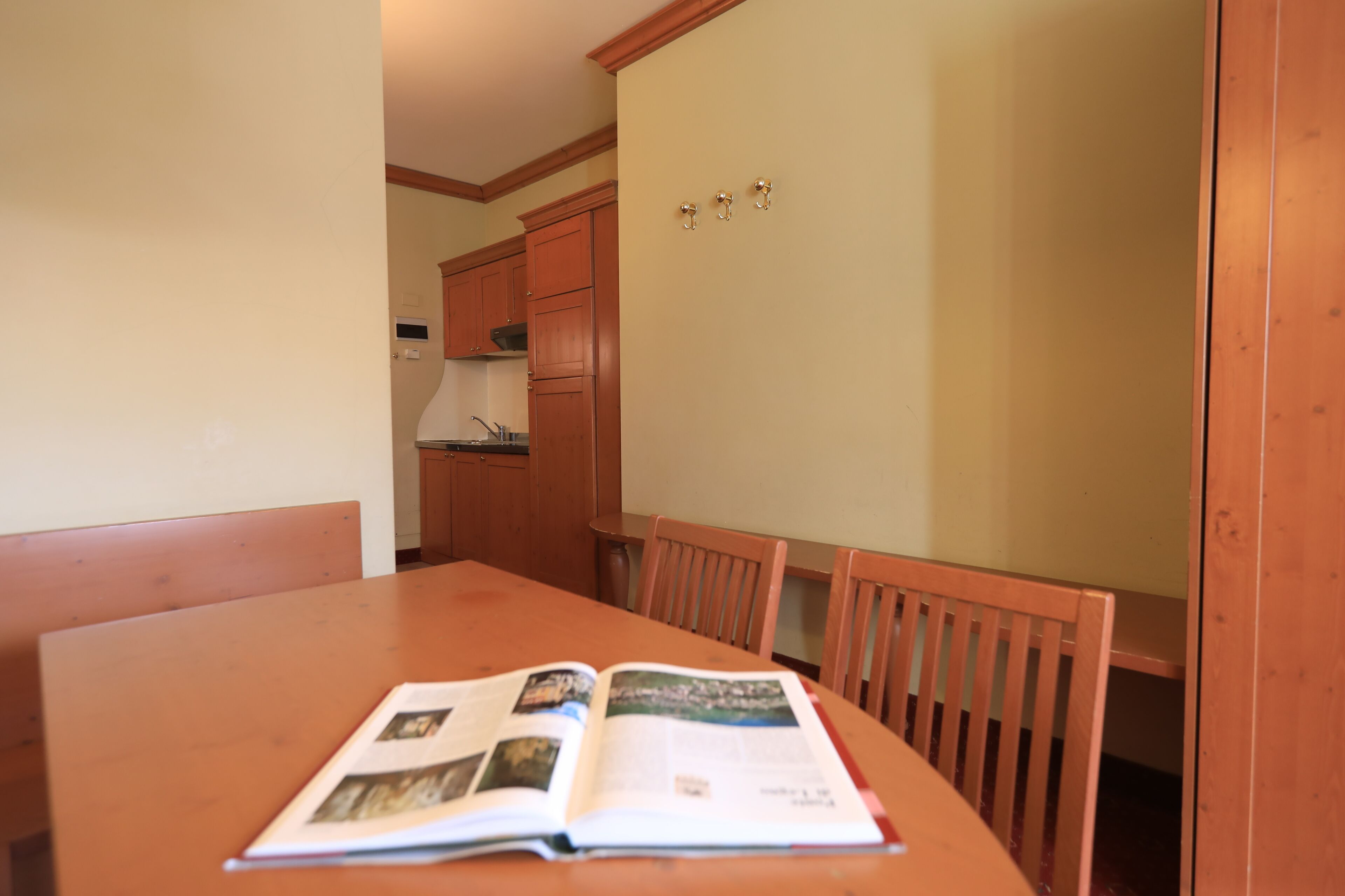 Un coin repas avec une table et des chaises en bois, un livre ouvert sur la table, et une cuisine en arrière-plan.