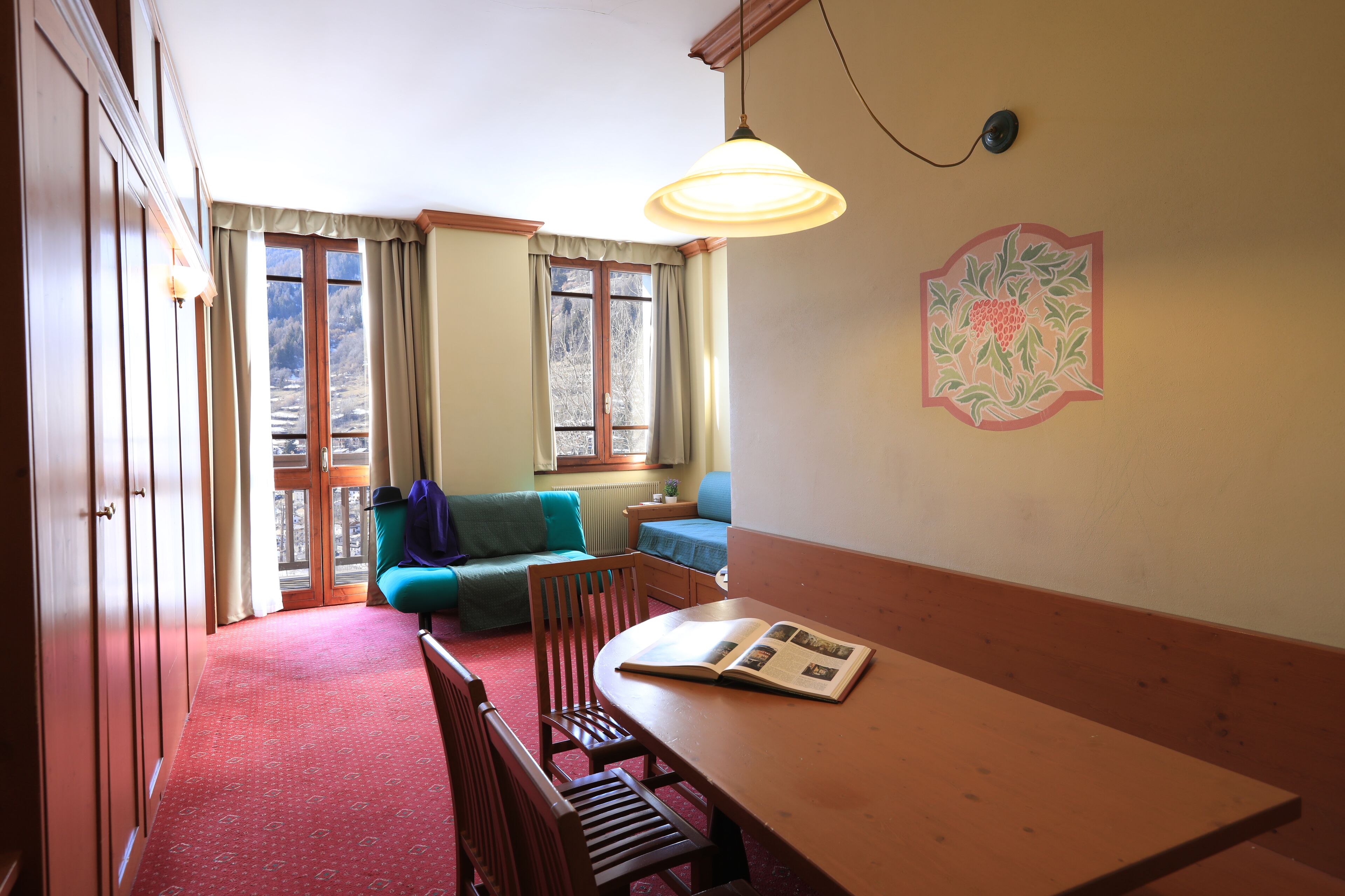 Een kamer met een houten tafel, stoelen, een bank, een raam met gordijnen, een hanglamp en een decoratie aan de muur.