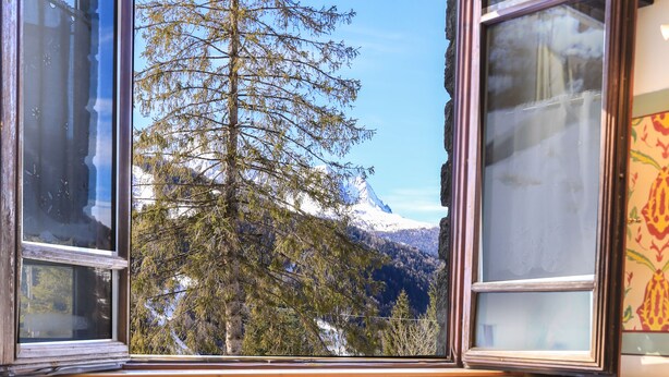 A table set for breakfast with a croissant, a cup of coffee, and a glass of orange juice. A jar of cookies is on the table, and a view of a mountain and trees is visible through the window.