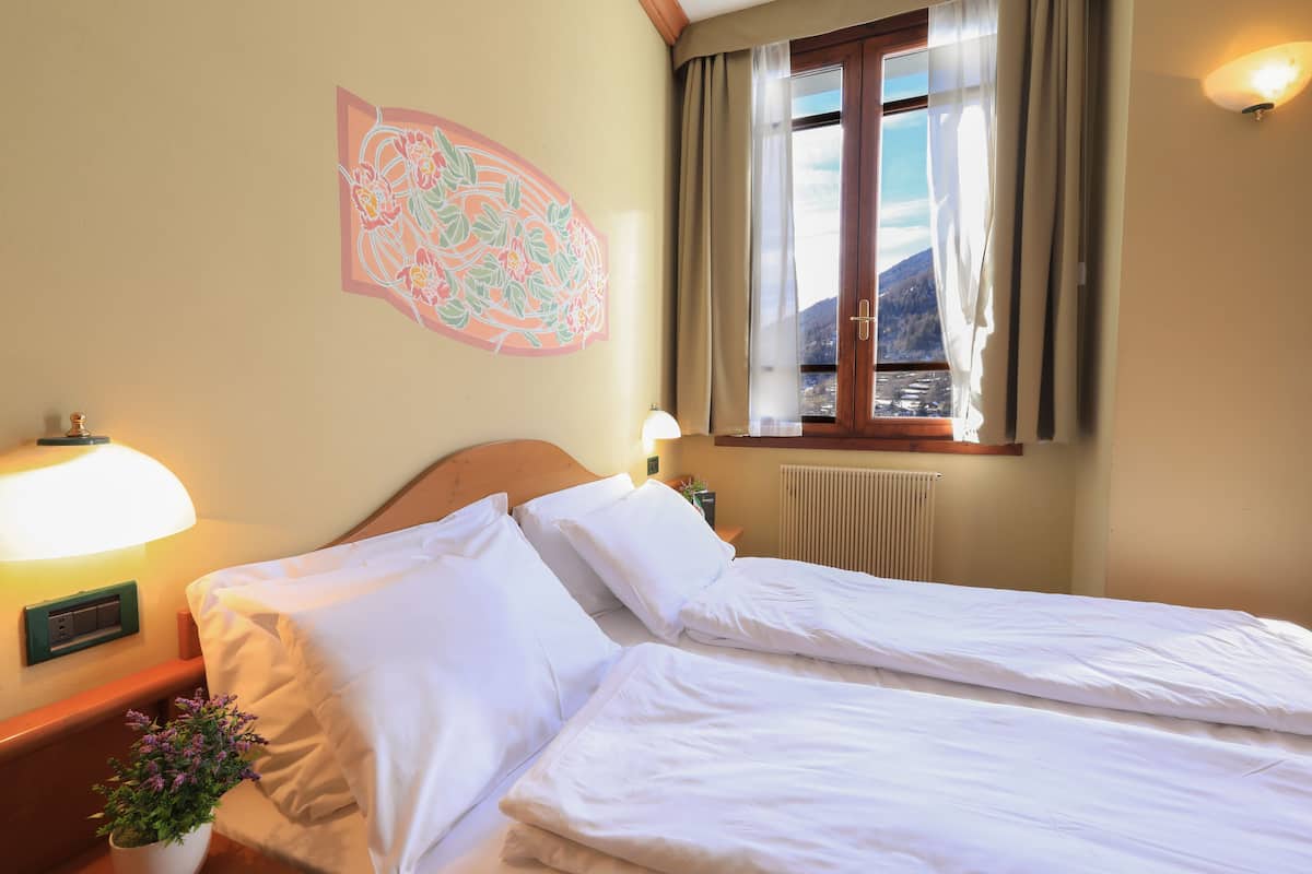 Un lit double avec du linge de lit blanc, une table de chevet avec une lampe, une plante en pot et une fenêtre avec des rideaux.