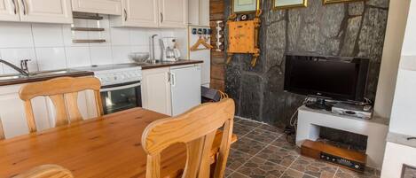 A kitchen with white cabinets, a wooden dining table, and chairs. There is a TV and a fireplace in the background.