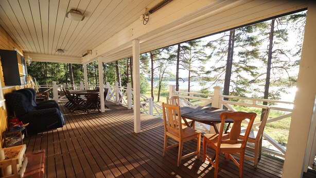 A covered outdoor area with wooden flooring, a table with chairs, and a view of a lake and trees. - Villa Helga (Kemiönsaari)