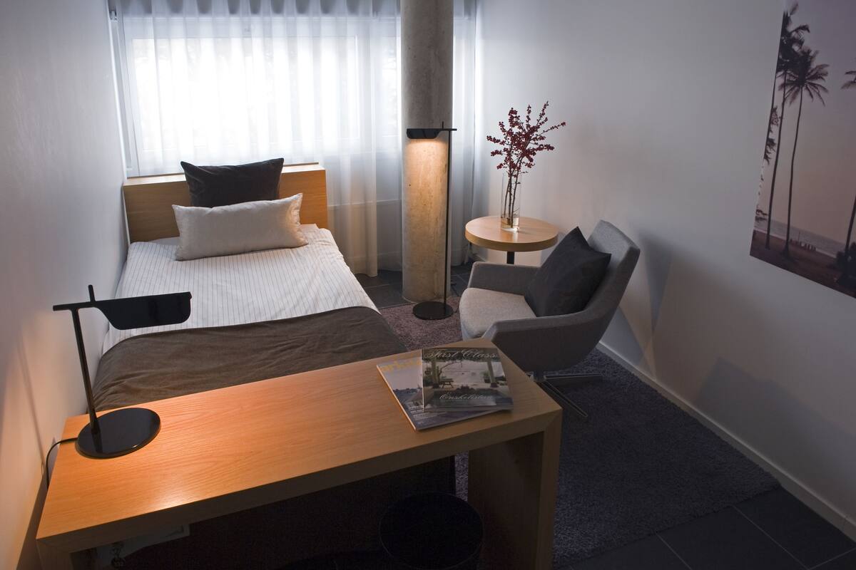 A hotel room with a bed, a desk, a chair, a small table, a lamp, and a picture on the wall.