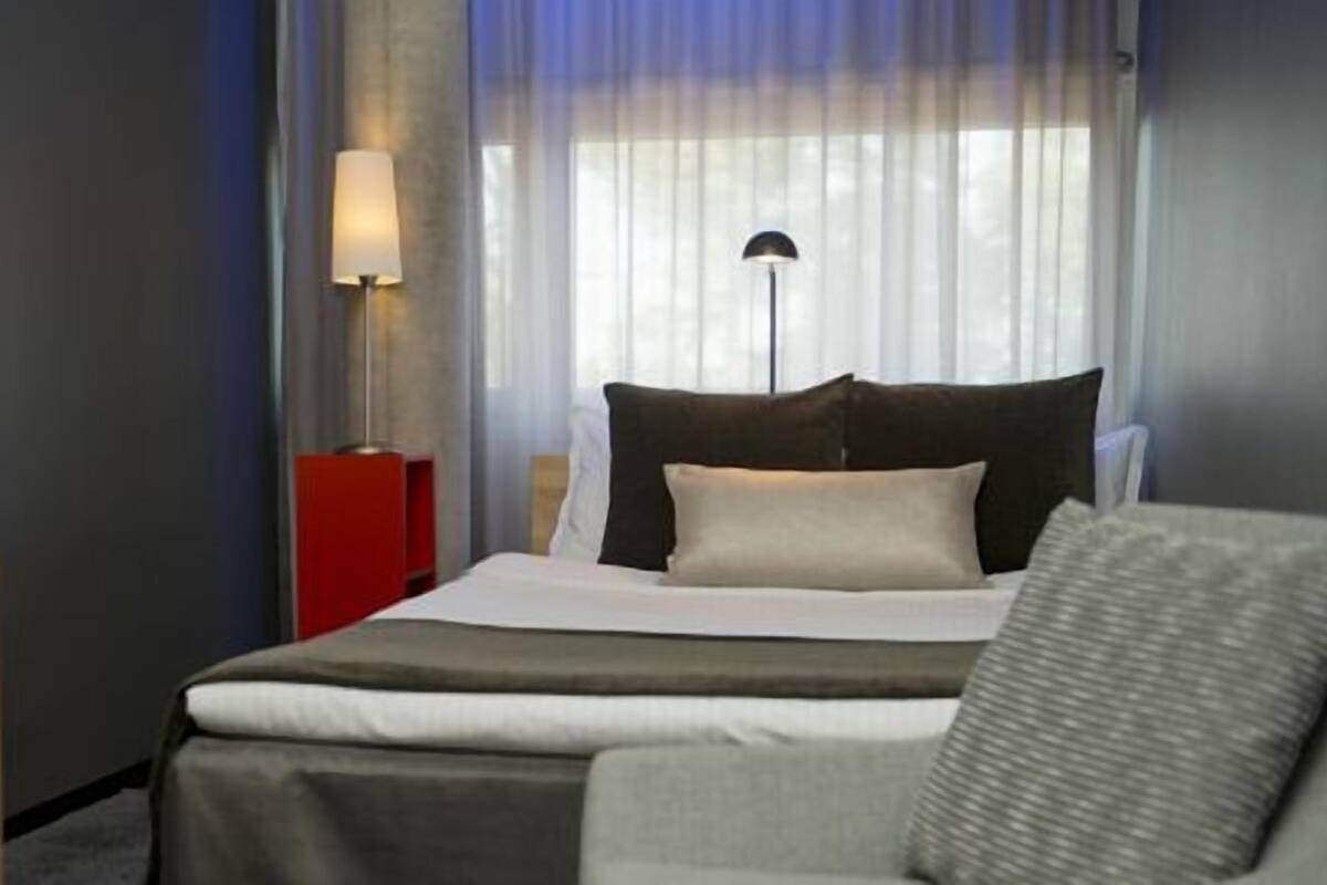 A hotel room with a bed, a nightstand, a lamp, and a view through sheer curtains.