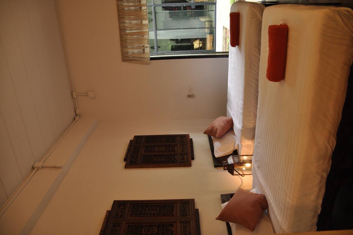 A small, narrow room with two beds, a window with blinds, and a wooden floor.