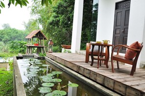 A wooden deck with a table and chairs, a small pavilion, and a pond with lily pads.