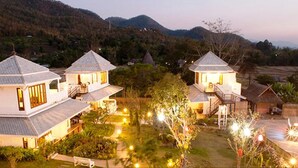 A cluster of white, two-story houses with thatched roofs, surrounded by lush greenery and terraced fields, set against a backdrop of hills. - Pai Vieng Fah (Pai)