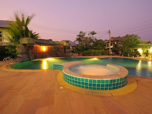 A swimming pool with a hot tub, surrounded by a wooden deck and a rock wall with a waterfall feature.