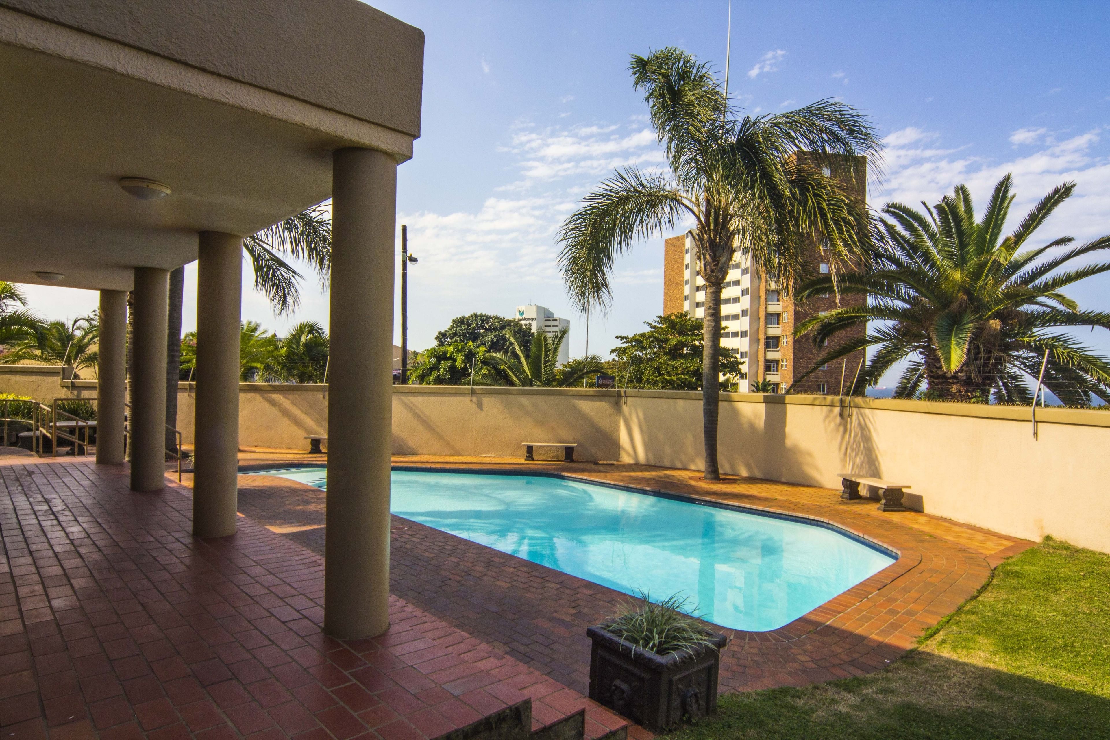Ein Schwimmbecken mit Pflasterterrasse, Palmen und Blick auf ein hohes Gebäude im Hintergrund.