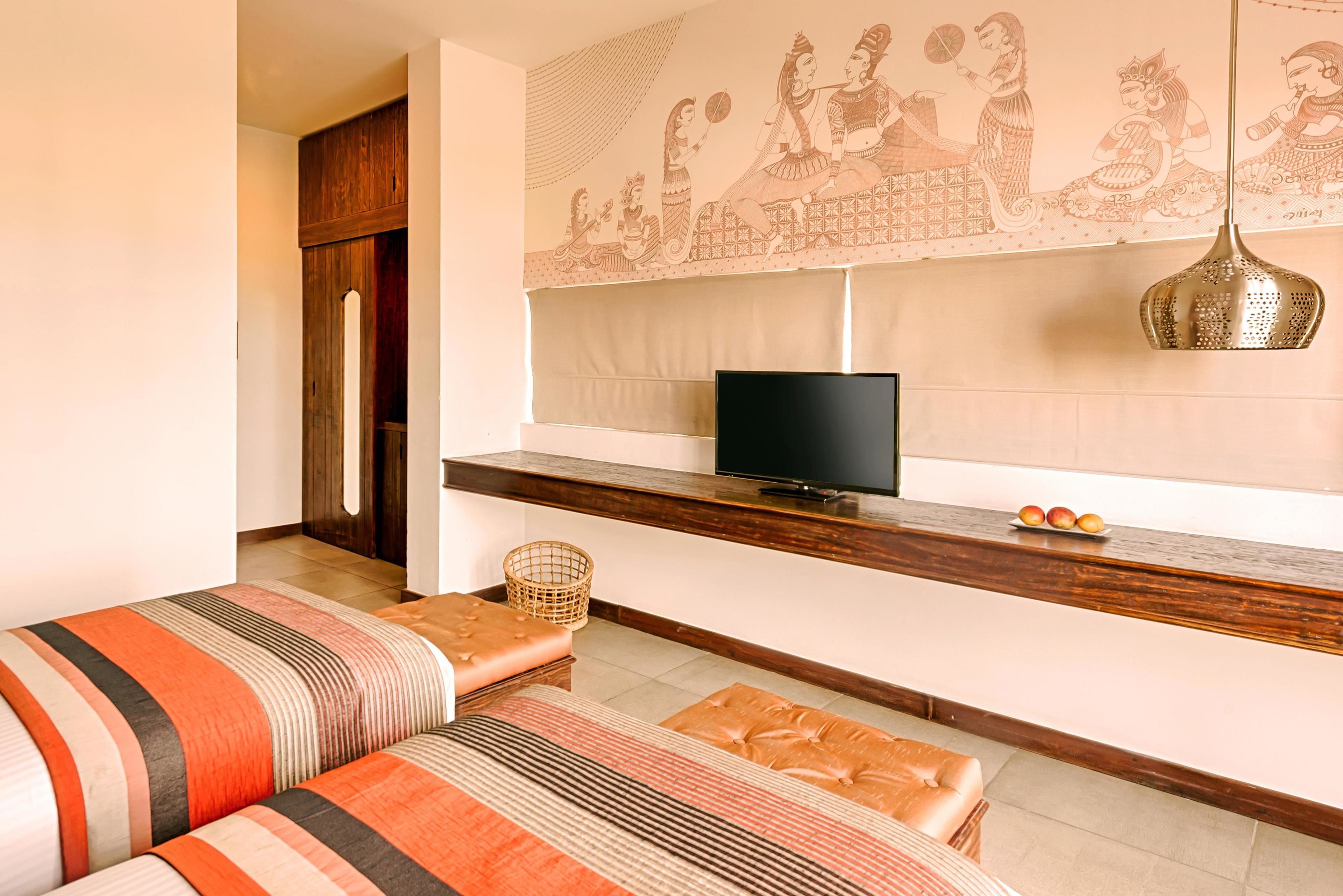 A modern hotel room with a TV, wooden bench, and a wall with traditional artwork.