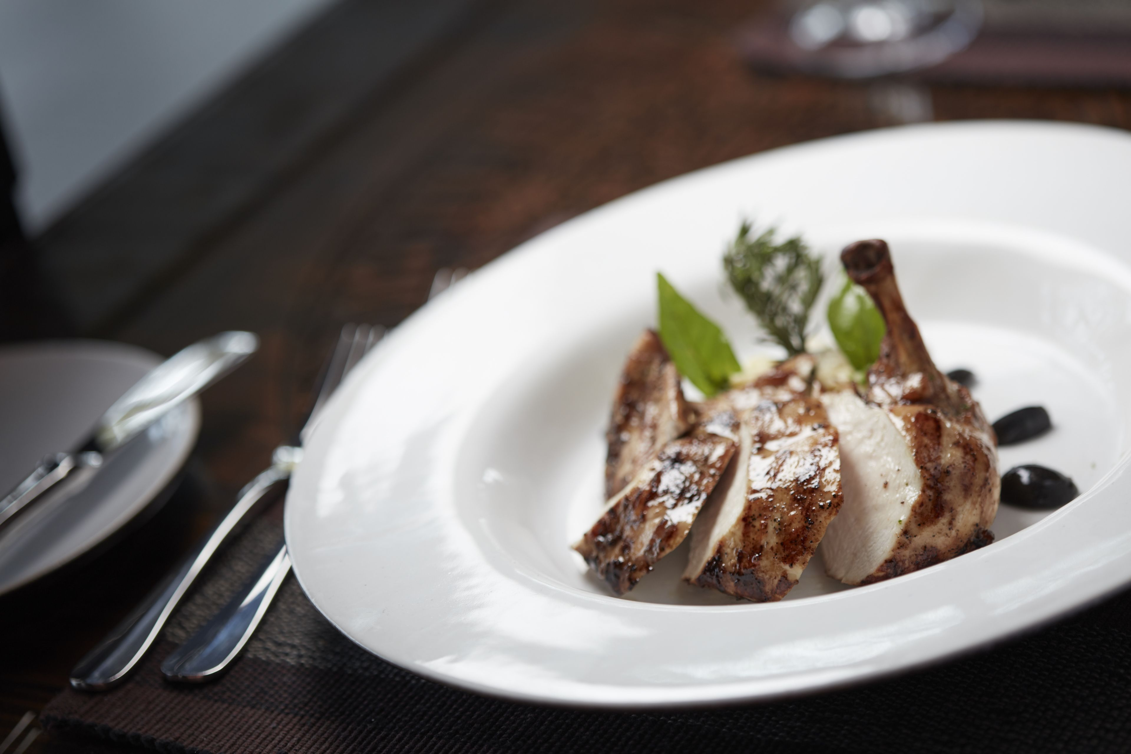 Une assiette blanche contenant un plat de poulet grillé, accompagnée d’une fourchette et d’un couteau posés sur une nappe sombre.