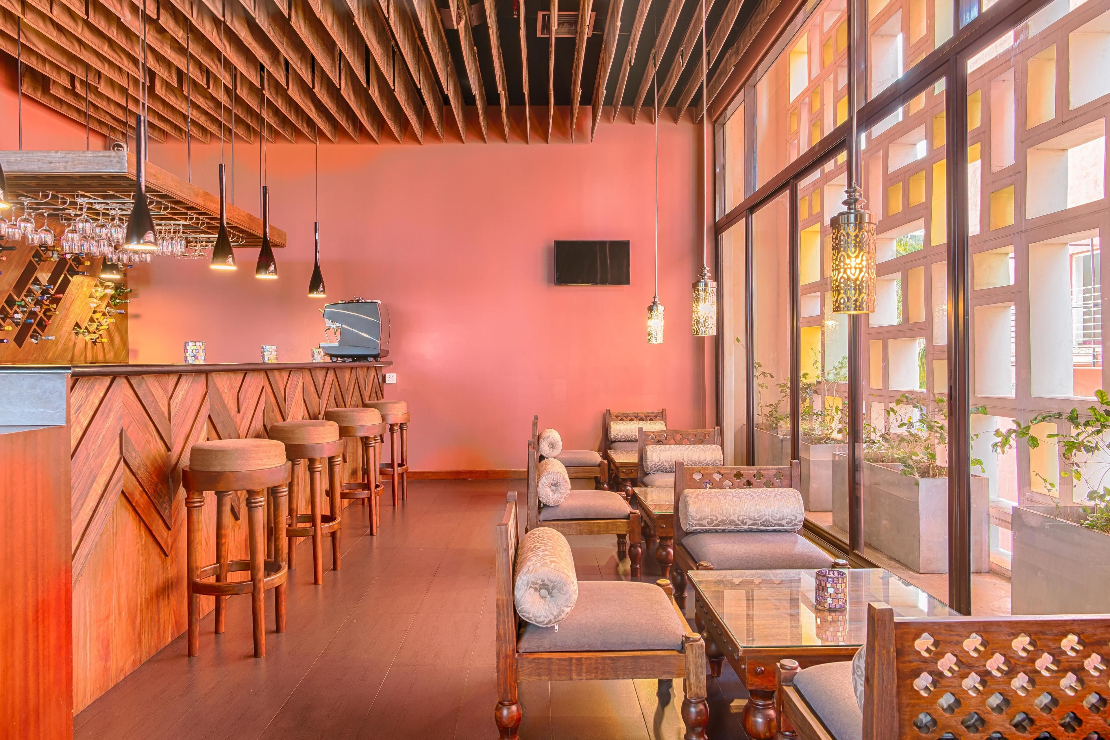A bar area with wooden stools, a bar counter, and hanging pendant lights. There are cushioned seating areas with patterned cushions and a large window with a view of greenery outside.