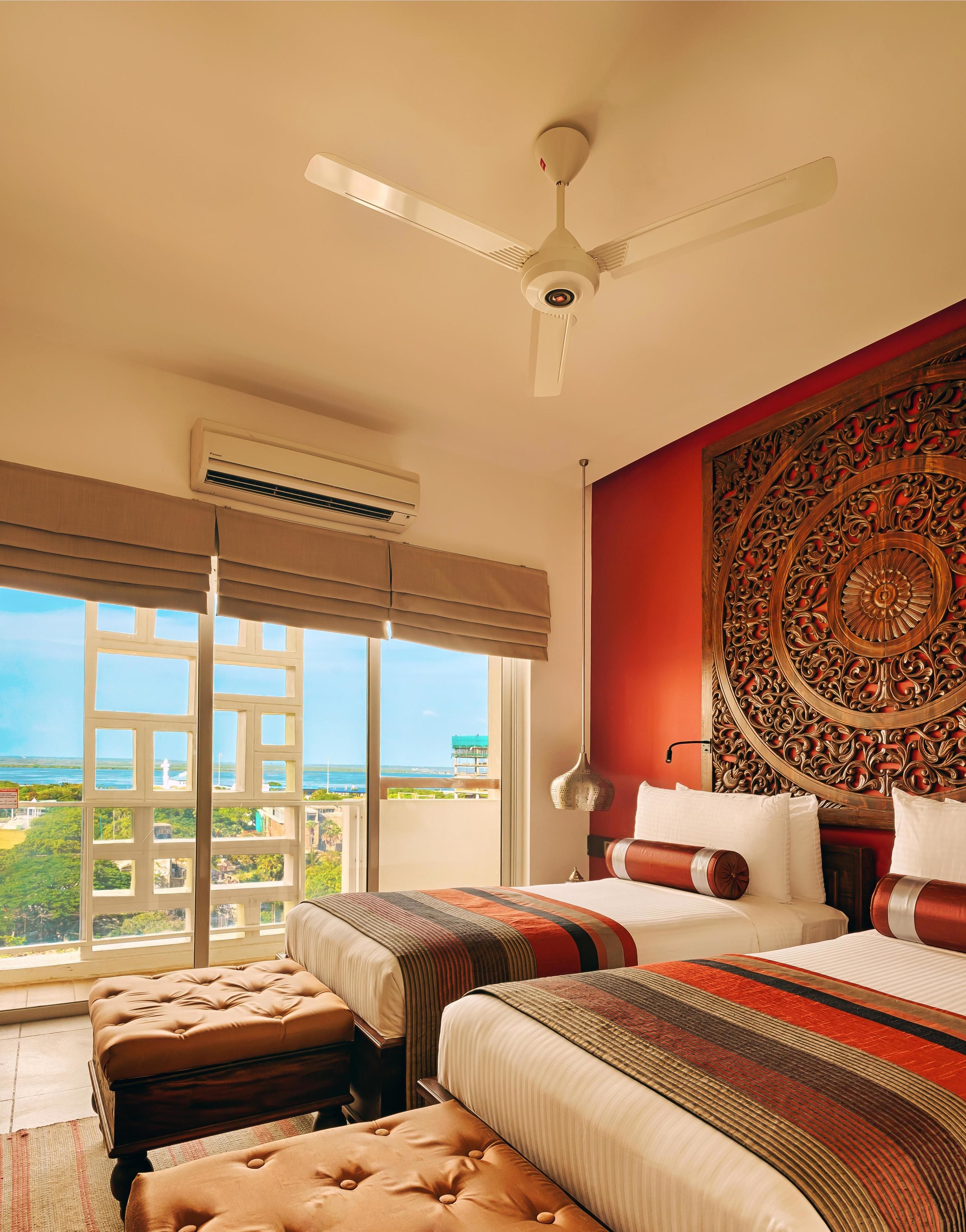 Quarto de hotel com cama grande, ventoinha de teto, ar condicionado, painel decorativo na parede e vista para a paisagem urbana pela janela.