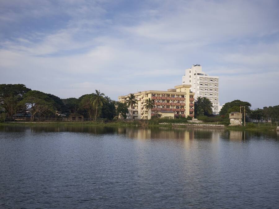 A building with multiple floors and balconies, situated near a body of water with trees and a clear sky in the background.