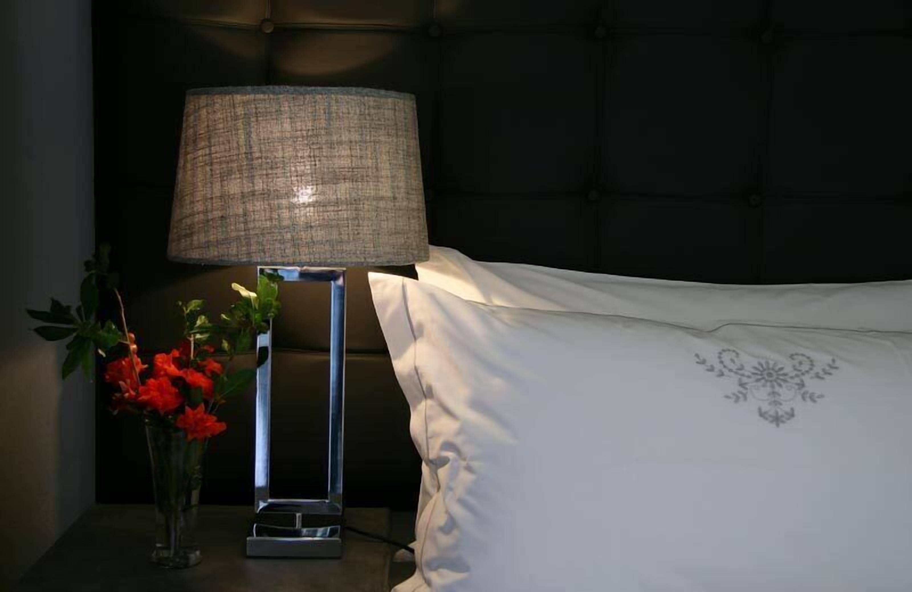 Een nachtkastje met een lamp, een vaas met rode bloemen en een wit kussen met een bloemendessin.
