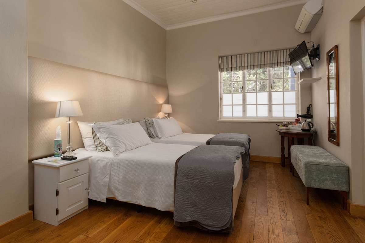 A bedroom with two beds, a bedside table with a lamp, a bench, a window with blinds, and a mirror on the wall.