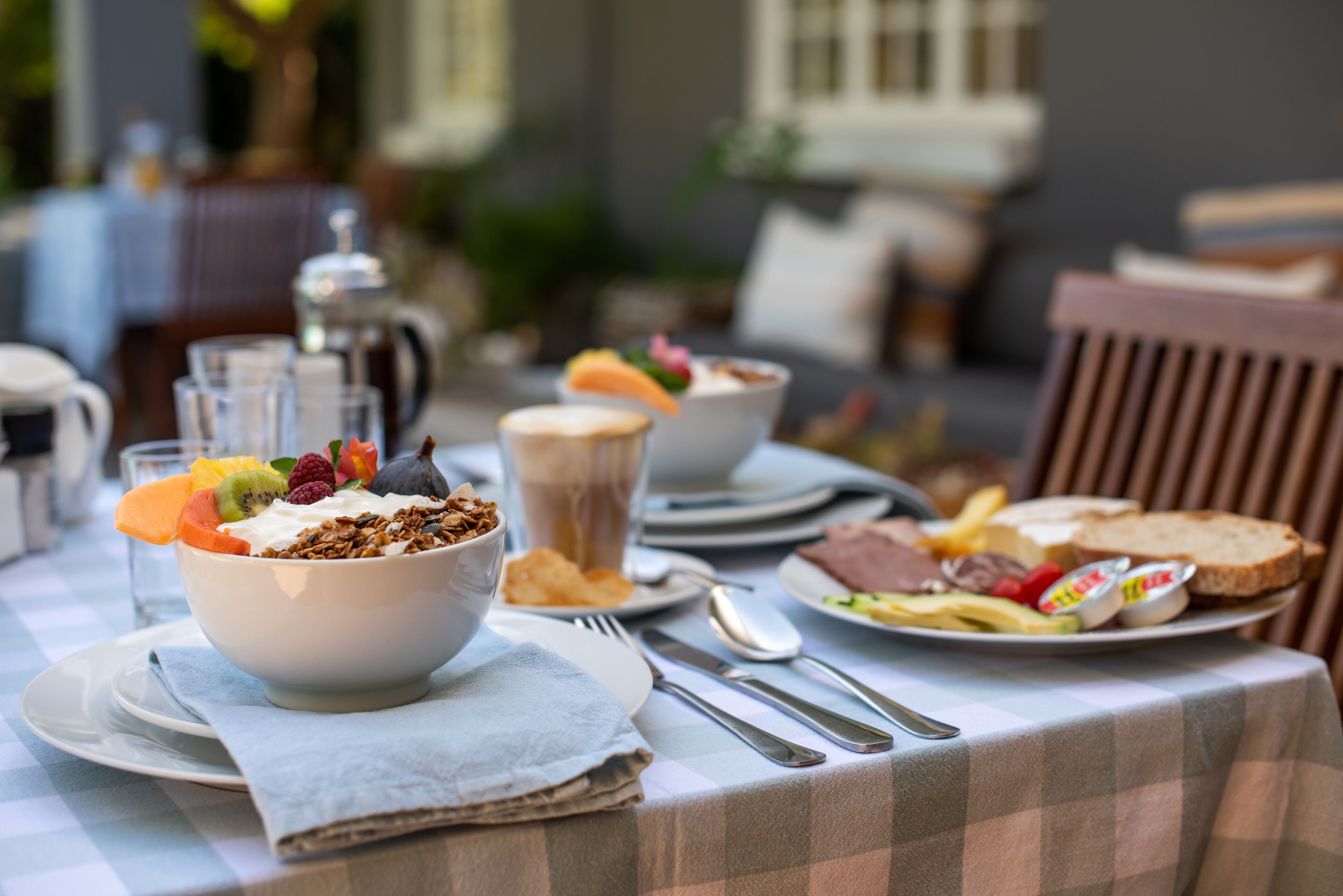 Ein Frühstückstisch mit kariertem Tuch, einer Schüssel mit Müsli und Früchten sowie Tellern mit Toast und Eiern.