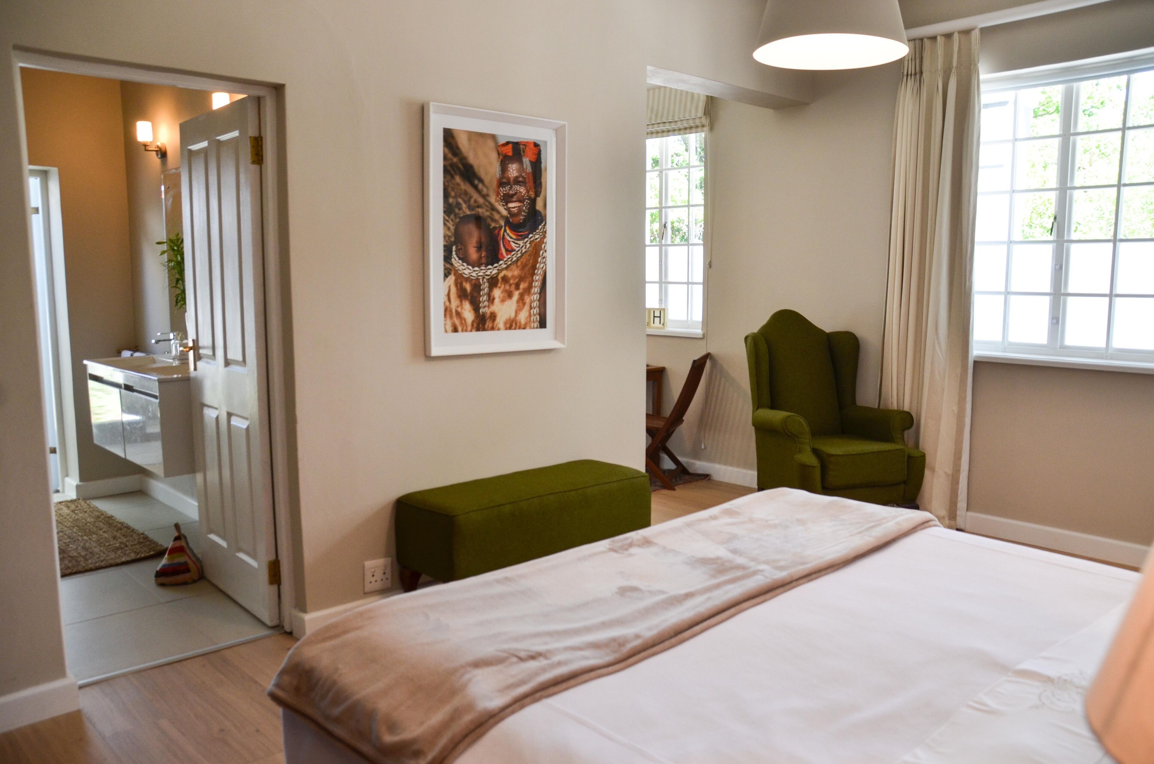A bedroom with a bed, a green armchair, a green ottoman, a framed picture on the wall, and a window with curtains.