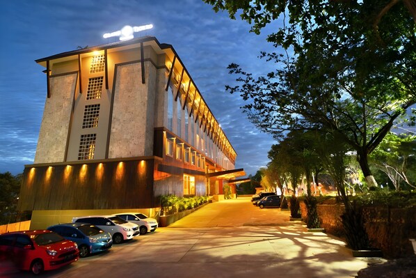 A modern hotel with a prominent entrance, a parking area with several cars, and a well-lit facade.