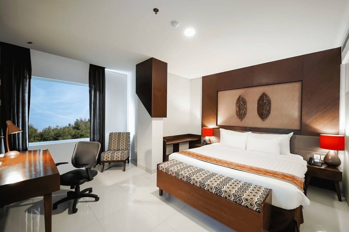 Ein modernes Hotelzimmer mit einem großen Bett, einem Schreibtisch, einem Sessel und Blick ins Freie.
