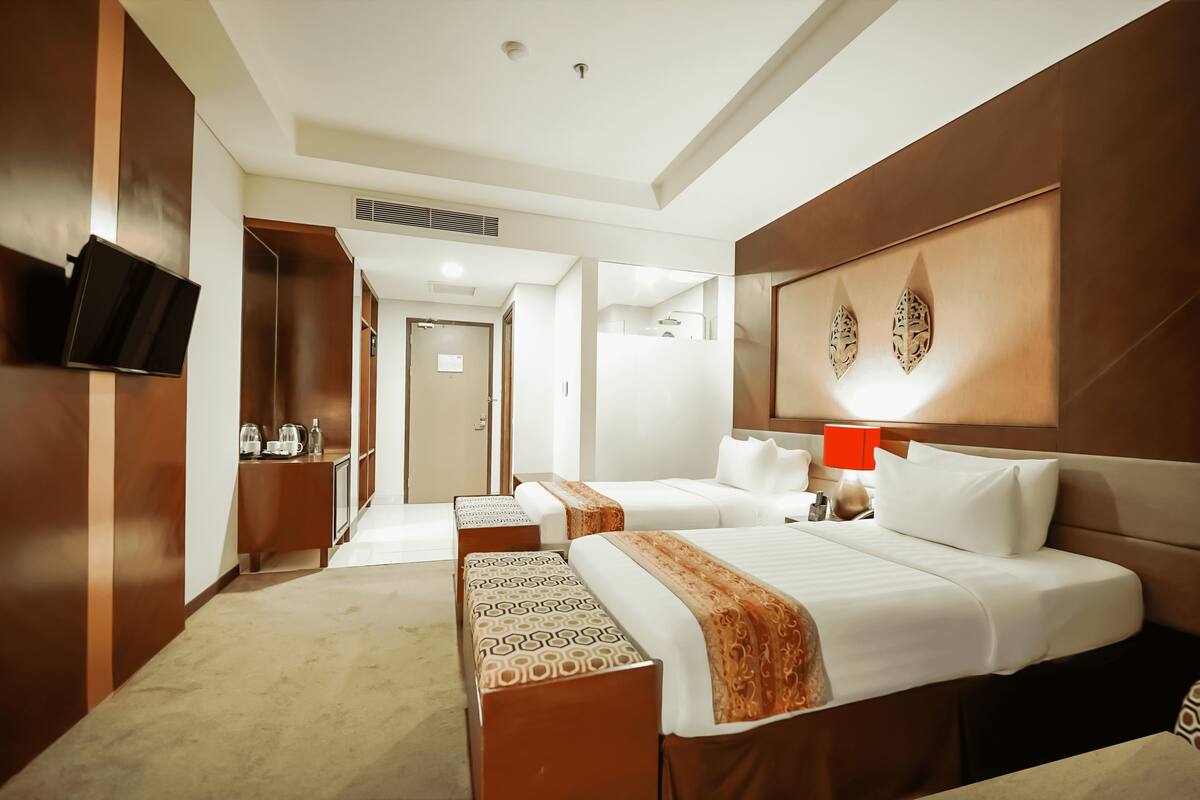 Ein Hotelzimmer mit zwei Betten, einem an der Wand montierten Fernseher, einem Schreibtisch mit Lampe und einem Spiegel.