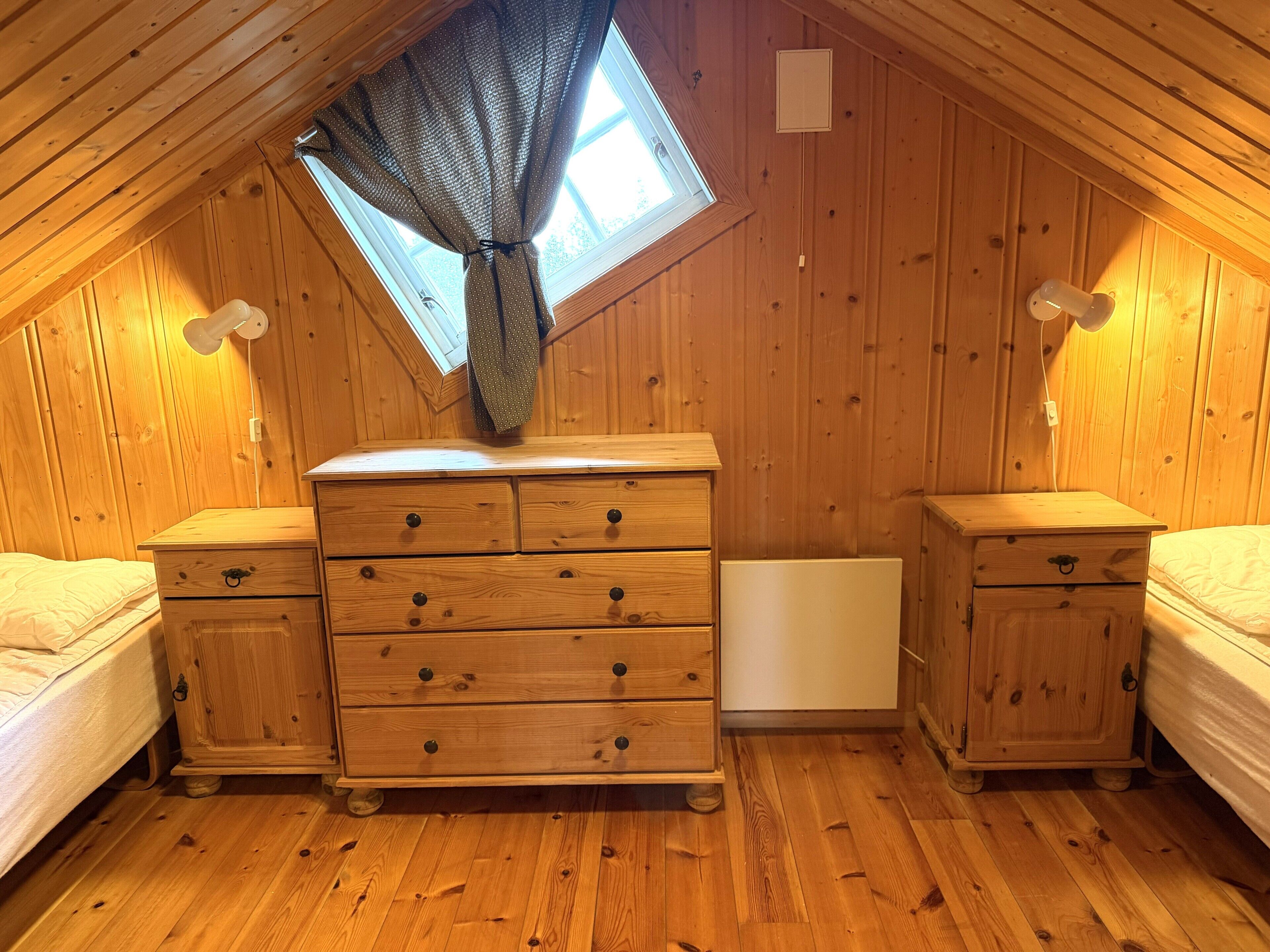 Una habitación acogedora con paredes de madera, un ventanal con cortina, dos mesitas de noche de madera, un aparador y una cama.
