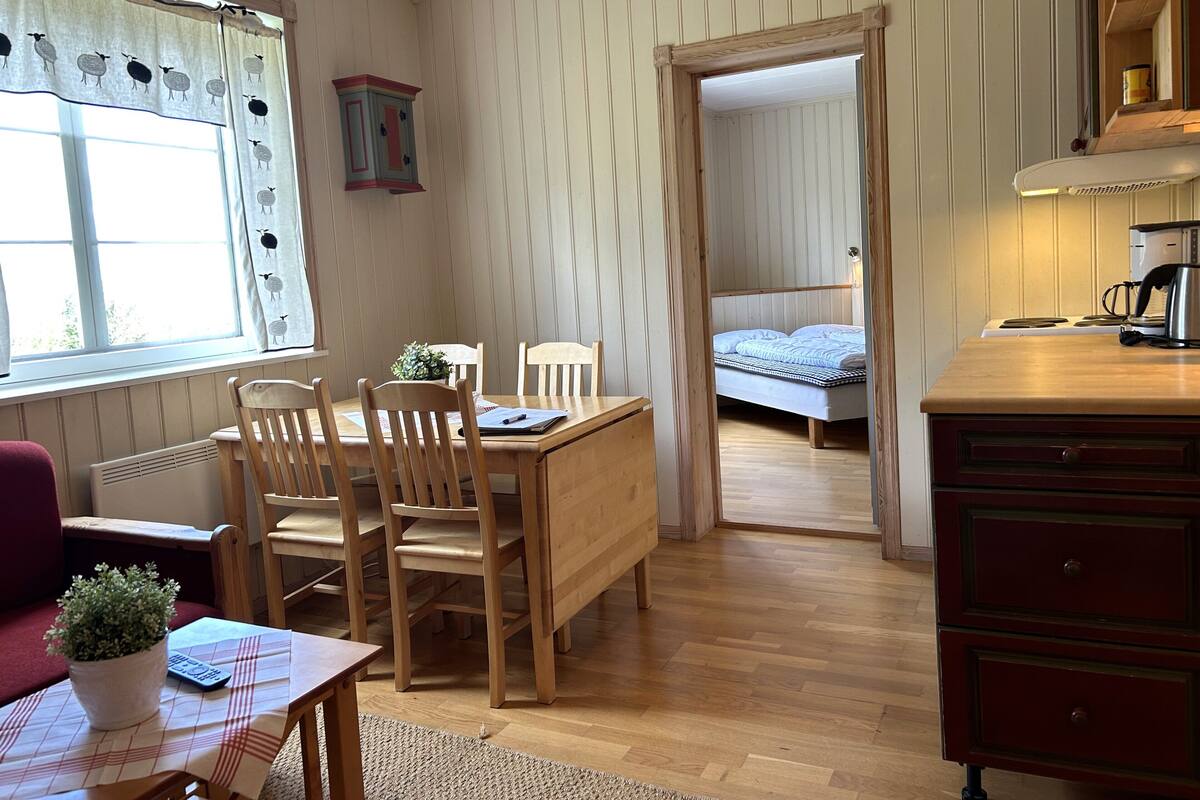 Ett mysigt rum med ett matbord i trä, stolar, ett litet skrivbord med en växt, en säng med ett blått täcke och en köksdel med en kaffebryggare.