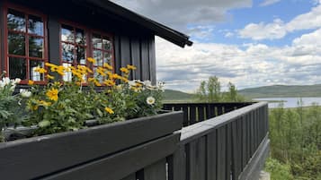 Un edificio de madera con balcón, un jardín con flores amarillas y blancas, y una vista de un lago y colinas al fondo.