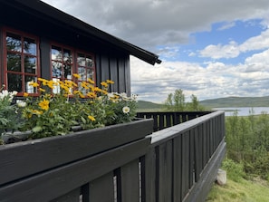A wooden building with a balcony, a flower bed with yellow and white flowers, and a view of a lake and hills in the background. - Torsetlia Fjellstue og Hyttegrend (Nore og Uvdal)