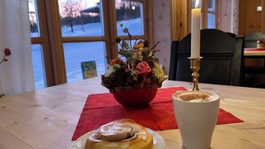 A wooden table with a plate of cinnamon roll, a white cup, and a floral arrangement with a candle. - Torsetlia Fjellstue og Hyttegrend (Nore og Uvdal)