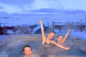 Uma banheira de hidromassagem com três pessoas, rodeada por uma paisagem nevada e uma casa ao fundo.