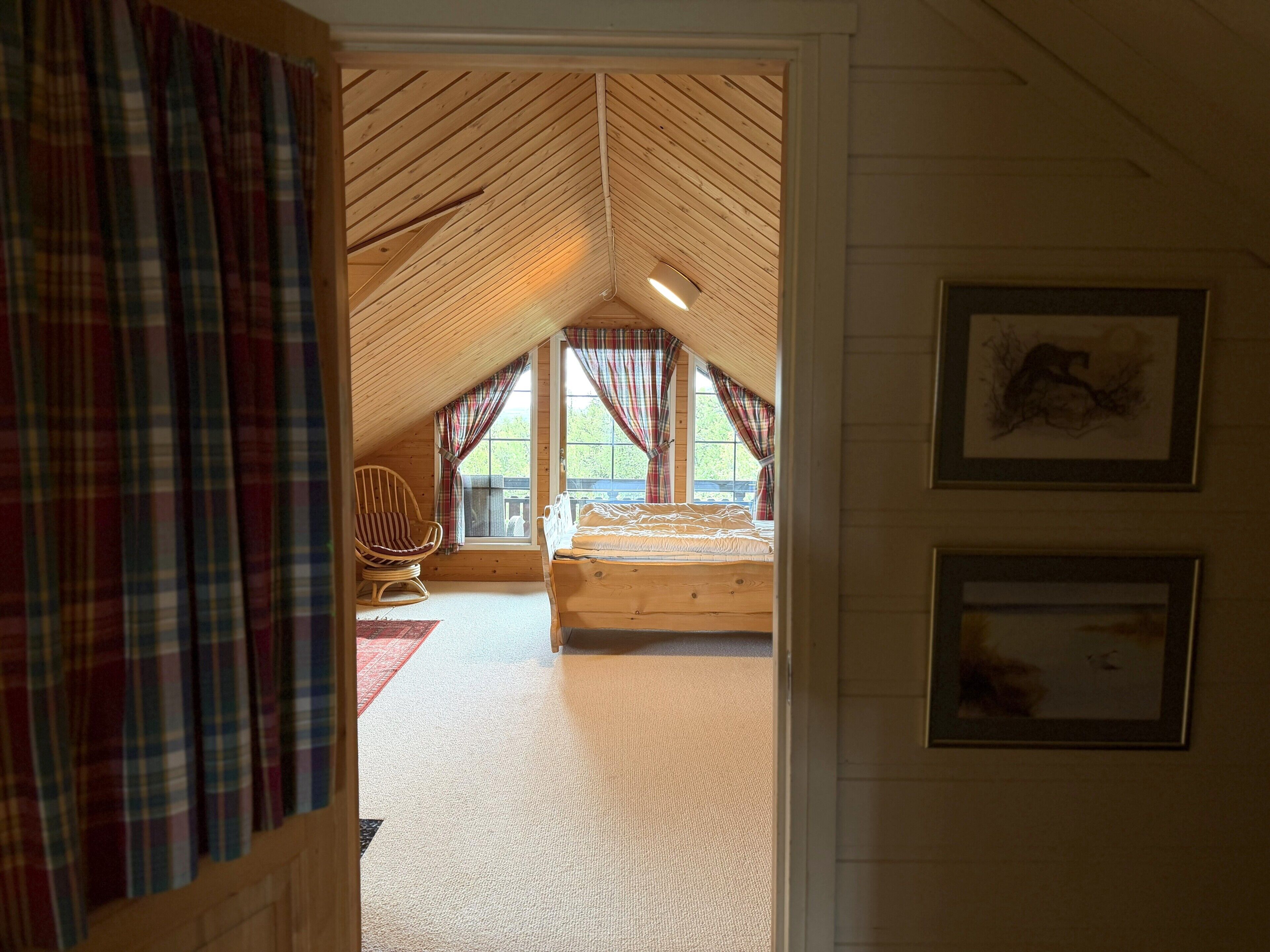 A room with a sloped ceiling, a bed, a chair, and two windows with curtains.