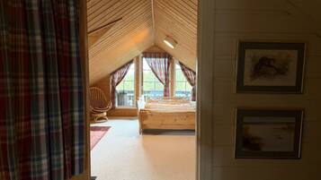A room with a sloped ceiling, a bed, a chair, and two windows with curtains.